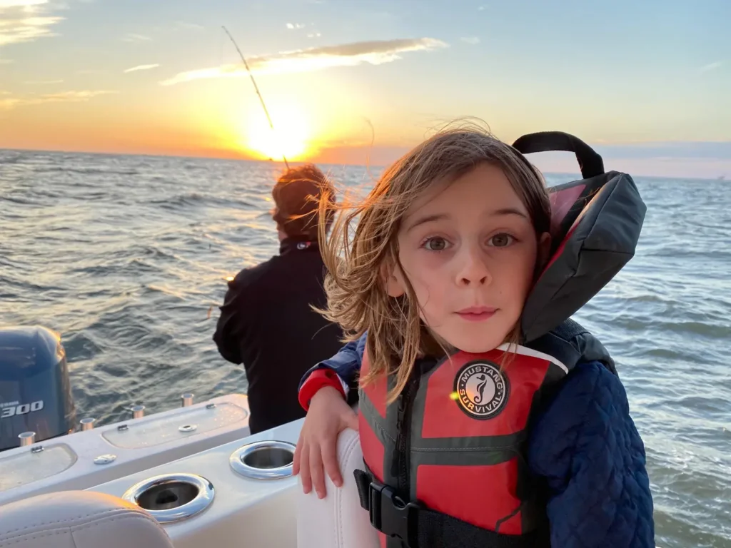 A child wearing a red life jacket stands aboard a fishing boat at sunset.