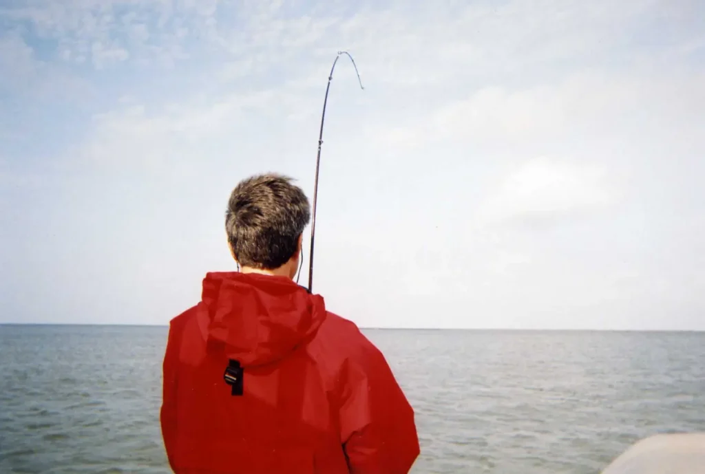 Rear view of an angler in a red jacket working a deeply bent fishing rod over open water.